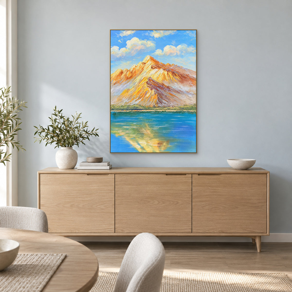 Blue lake and warm mountain ridges in a champagne-tone frame above a pale oak-tone sideboard