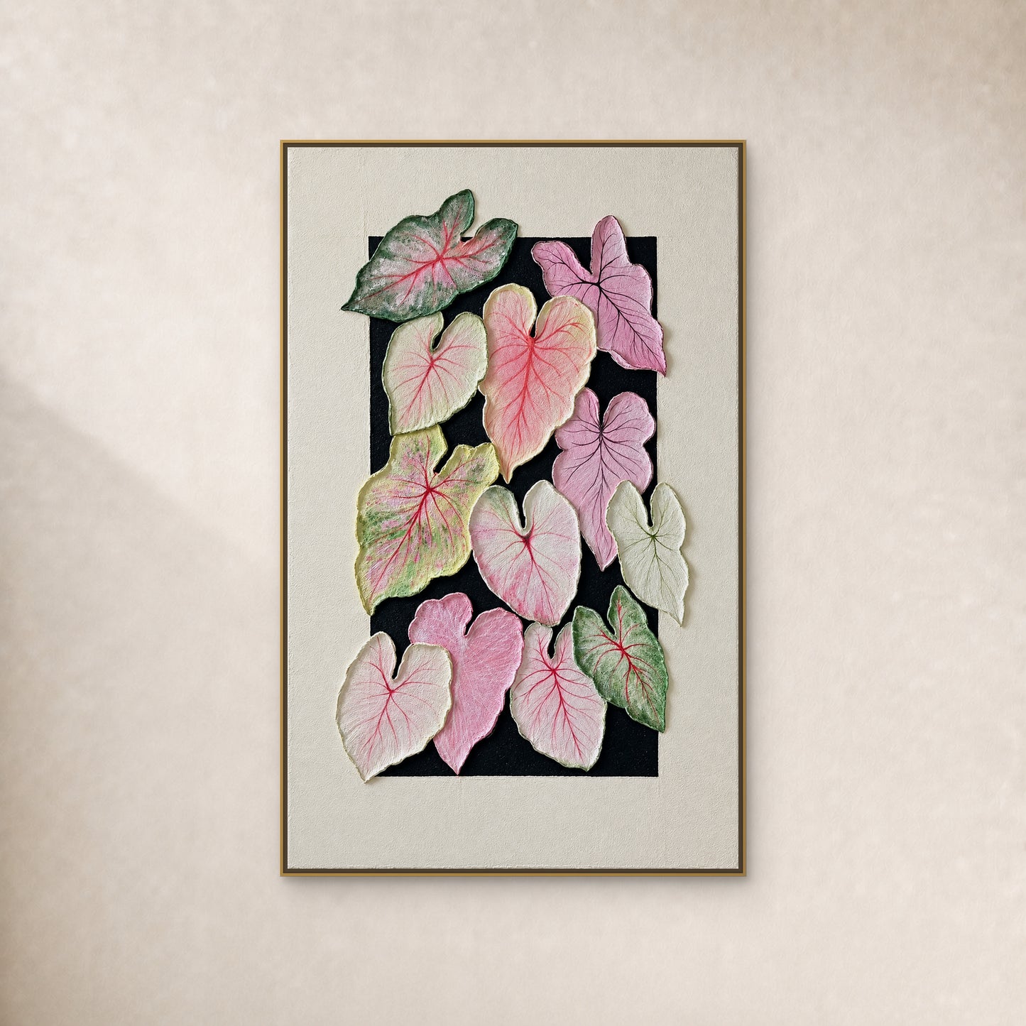 Twelve pink, lilac and green caladium leaves on a black painted field with an ivory surround, in a thin gold-tone floating frame on an ivory wall