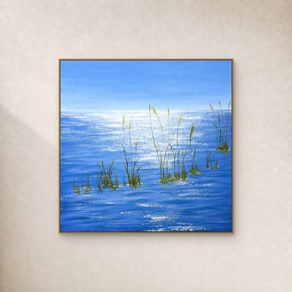 Square painting of blue water, white reflections and green grasses in a thin gold-tone floating frame on an ivory wall