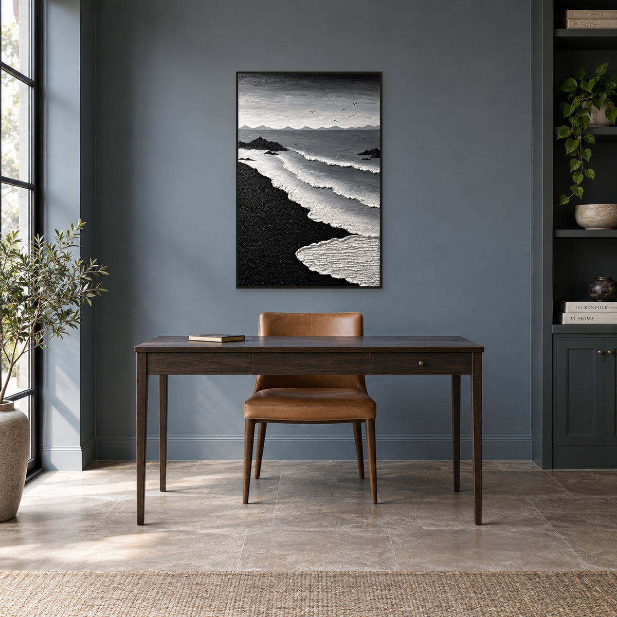Vertical black-sand seascape in a black floating frame above a dark wood desk and tan chair against a slate-grey wall