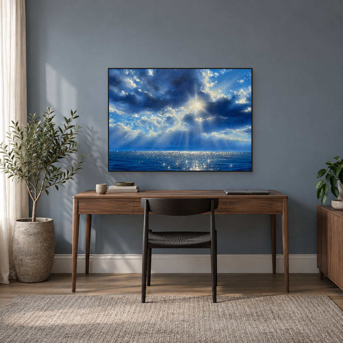Sunlit blue ocean painting in a thin black frame above a walnut desk against a blue-grey wall.