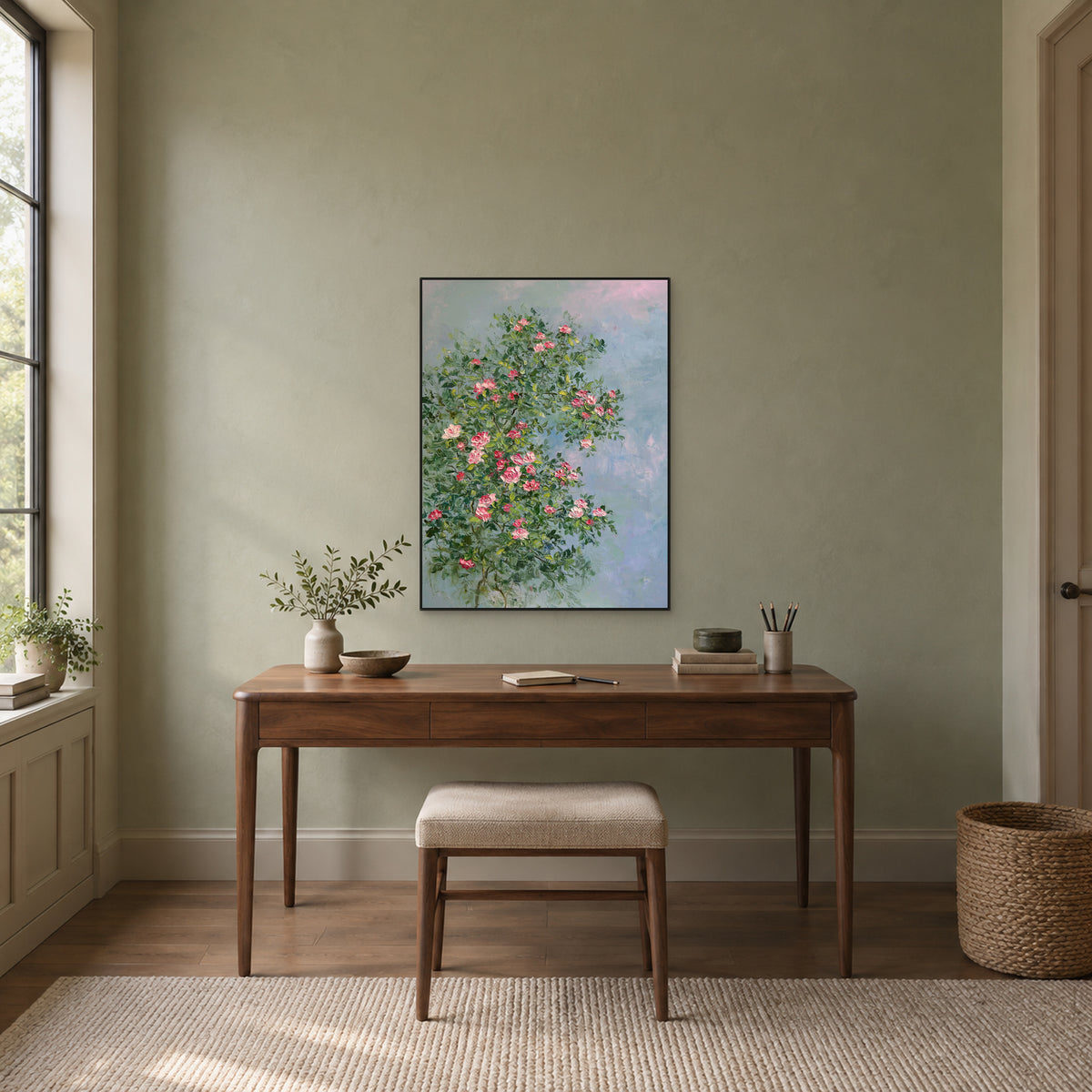Pink rose and blue-grey floral painting in a thin black frame centred above a wooden desk