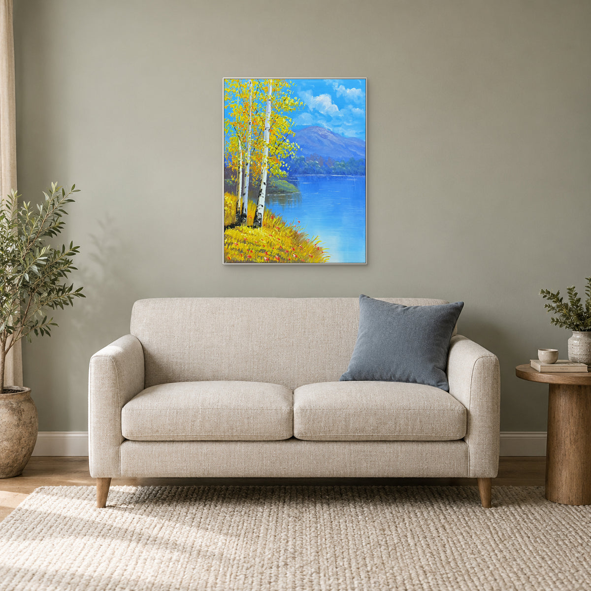 White-framed birch lake landscape above a small cream sofa, with a blue cushion and plants.