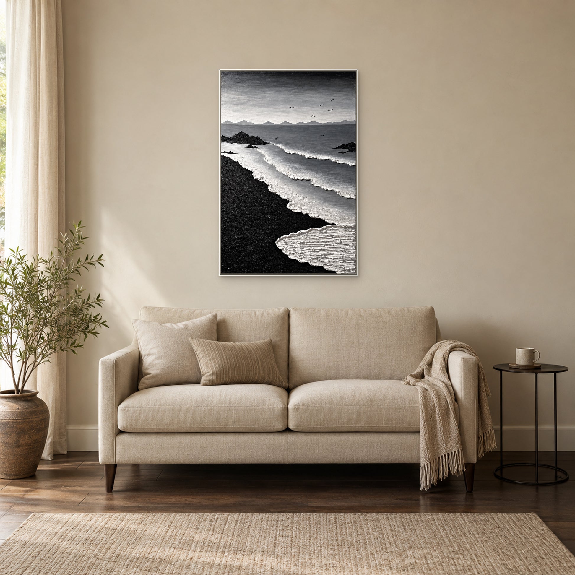 Textured white surf and black sand in a white floating frame above an oatmeal settee beside a window and a small black table