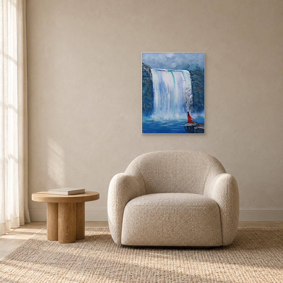 White-framed blue waterfall painting above a rounded oatmeal armchair, beside a low oak table in a reading corner.