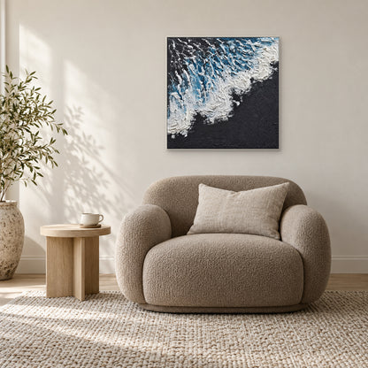Diagonal white surf painting in a white frame above a taupe armchair beside a small round wooden table