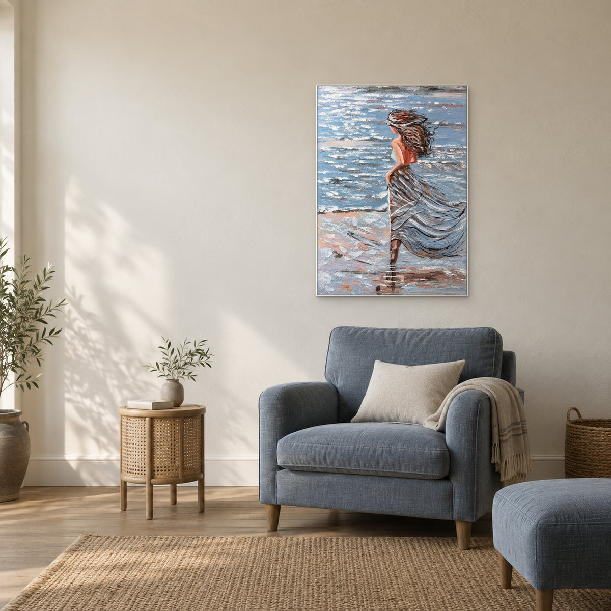 Vertical coastal portrait in a white floating frame above a slate-blue armchair beside a round woven side table