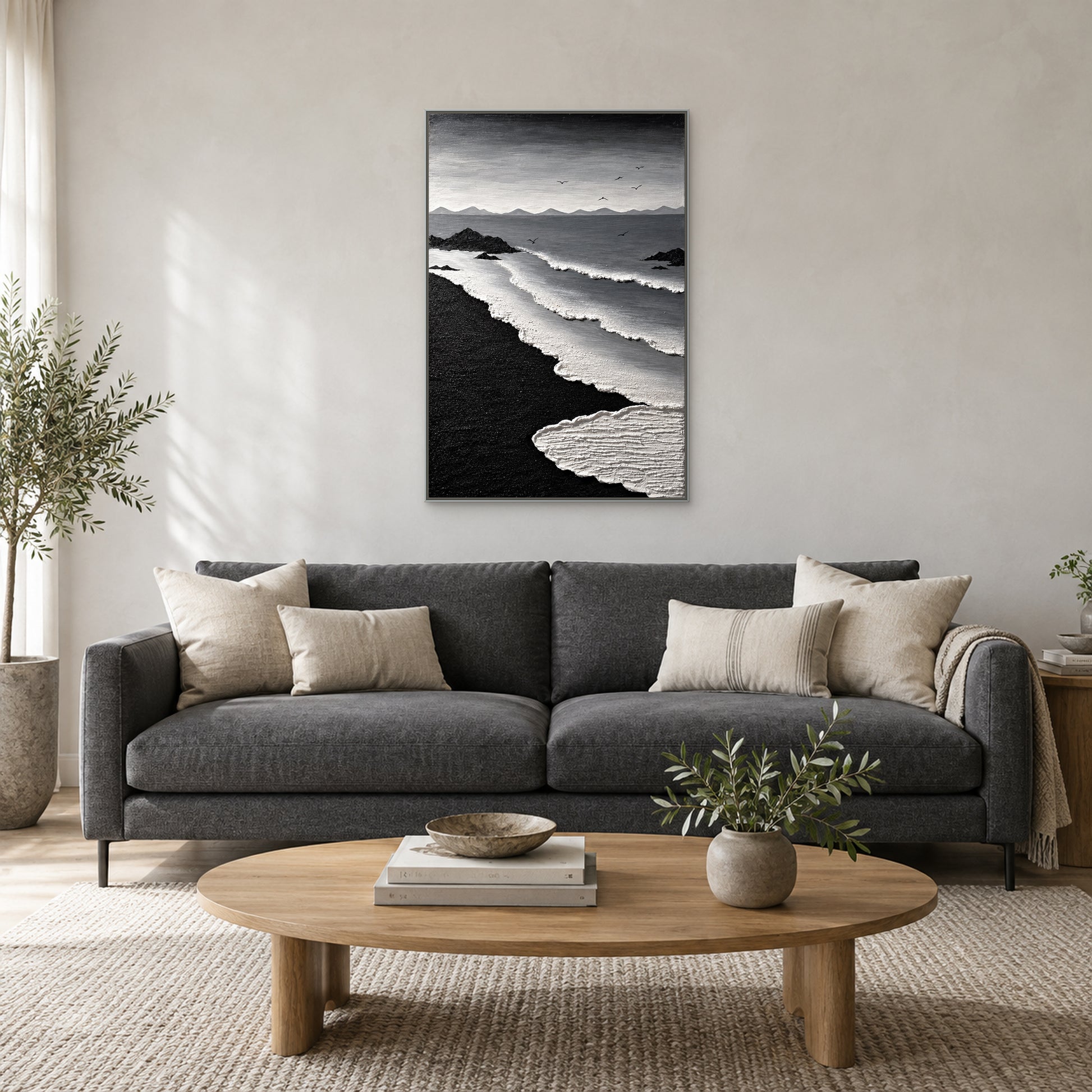 Monochrome coastal painting in a silver-tone floating frame centred above a charcoal sofa with ivory cushions and an oval wood coffee table
