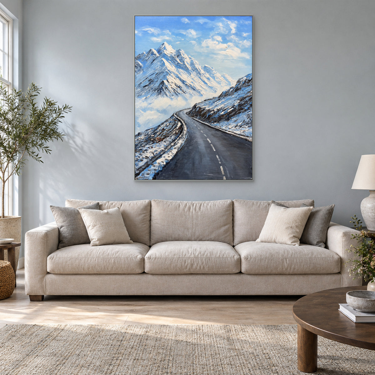 Snow peaks and a winding road in a silver-tone floating frame above a cream sofa against a blue-grey wall
