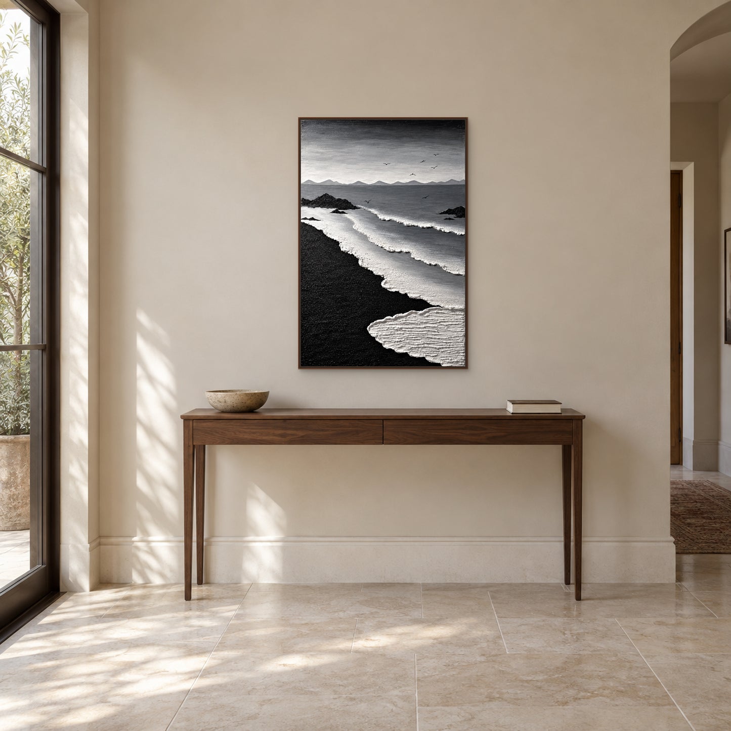 Black sand and white surf painting in a walnut-tone floating frame above a slim wood entryway console with a low bowl and books