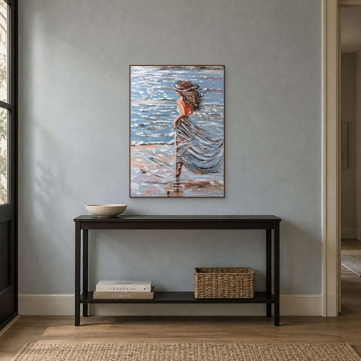 Blue-grey coastal woman painting in a walnut-toned floating frame above a slim black console against a blue-grey wall