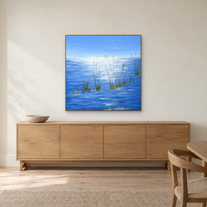 Blue water and delicate grasses in a champagne-tone frame centred above an oak-toned sideboard