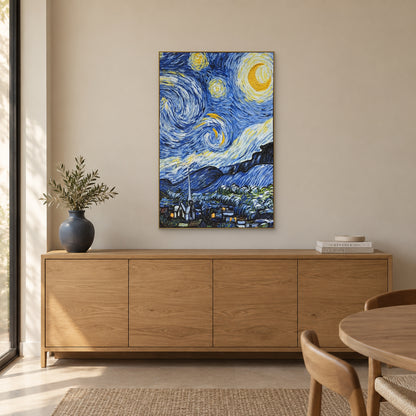 Starry Village in a champagne-tone frame above an oak-look sideboard with a blue-grey vase, leafy branches and books.