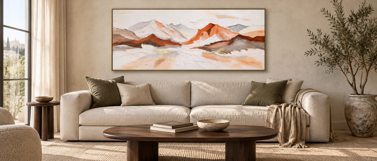 Copper and ivory mountain painting in a gold frame above a linen sofa, with a walnut coffee table, woven rug and soft window light.