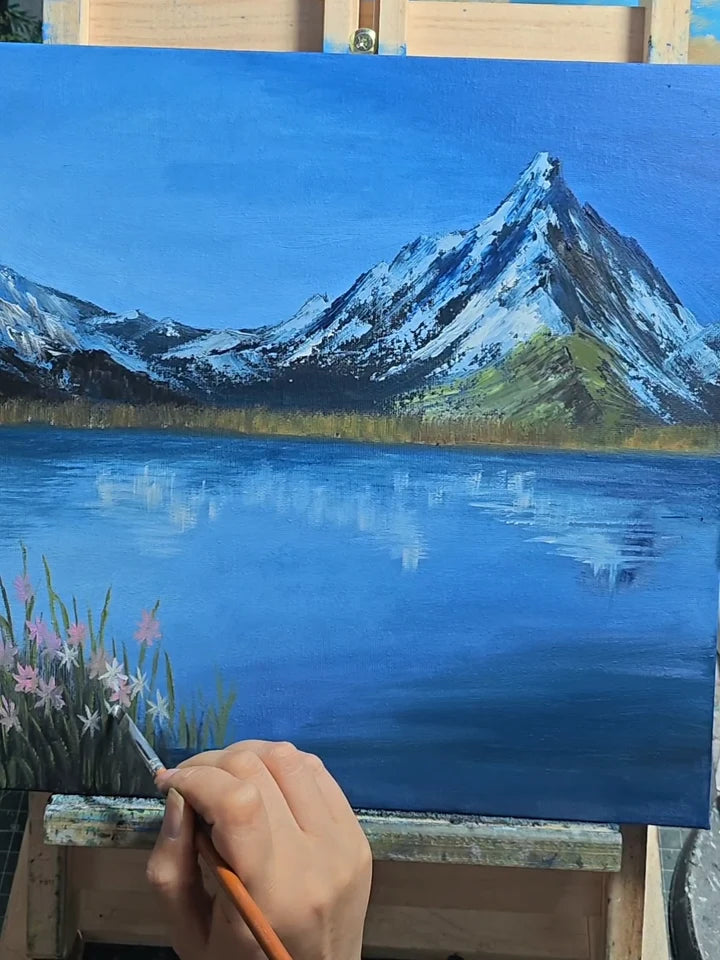 An artist painting flowers beside a blue alpine lake and mountain landscape