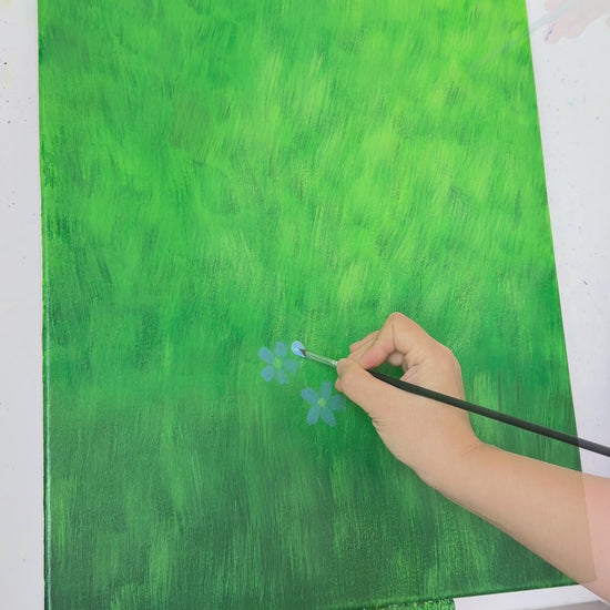 A hand paints small blue flax flowers across a green canvas, followed by the completed floral painting