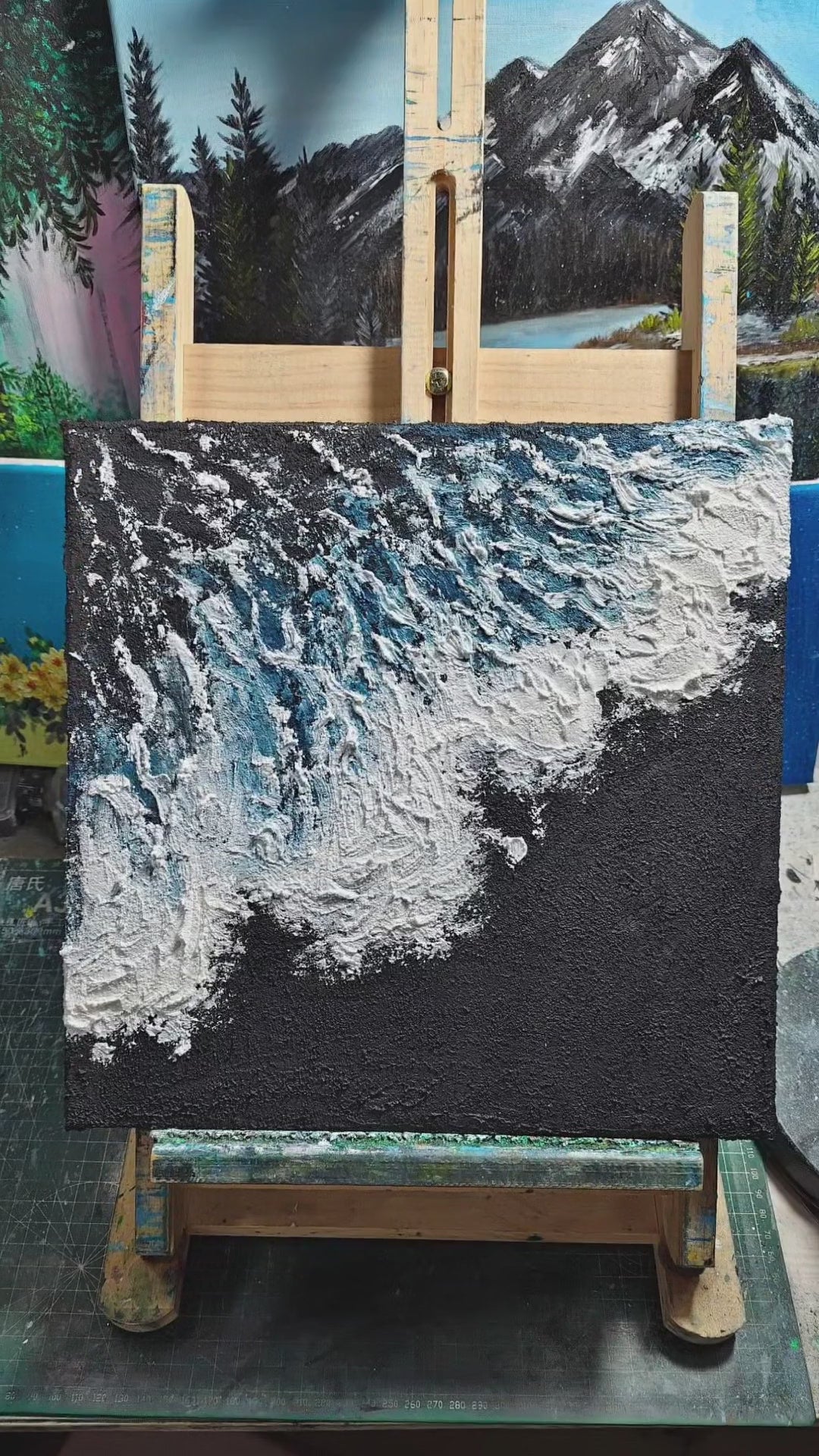 A square blue wave painting shown on an easel, then created through charcoal ground, white foam and fine blue brush marks